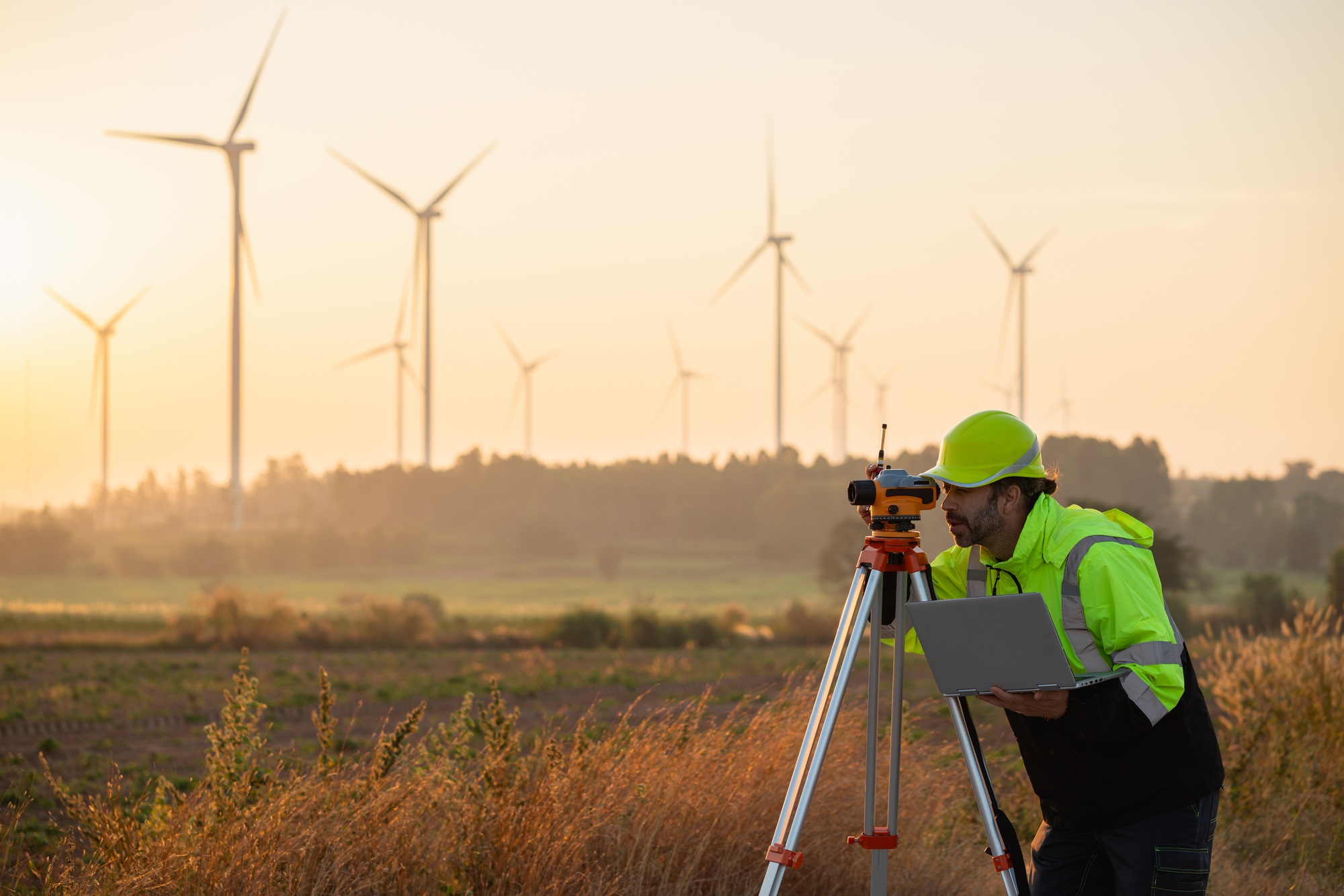At sunset, a field engineer surveys land as wind turbines turn—where tech meets clean energy.